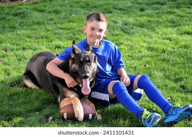 Boy wearing blue and his German Shepherd football playing dog taking a rest on the grass in backyard after running and playing football together. Boy has arm around dog and dog has football und er paw - Powered by Shutterstock