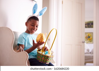 Boy Wearing Bunny Ears Sitting On Seat Holding Chocolate Egg He Has Found On Easter Egg Hunt