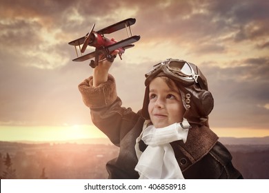 Boy wearing old-fashioned aviator hat, scarf and goggles holding a wooden biplane up in the air with sunset