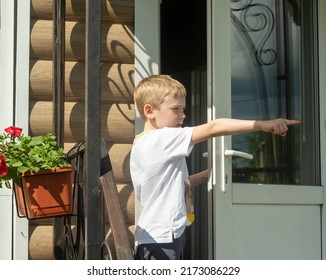 A Boy In A White T-shirt With Blond Hair Looks Out From Behind The Front Door Of A Country House And Points To The Side. Close-up.