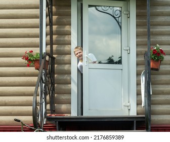 A Boy In A White T-shirt With Blond Hair Looks Out From Behind The Front Door Of A Country House. Copy Space.