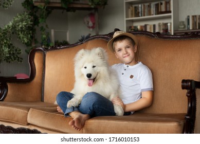 A Boy In A White T-shirt, Blue Jeans And A Wicker Hat Sits On A Sofa With His Bare Legs Crossed And Hugs A White Fluffy Samoyed Dog.