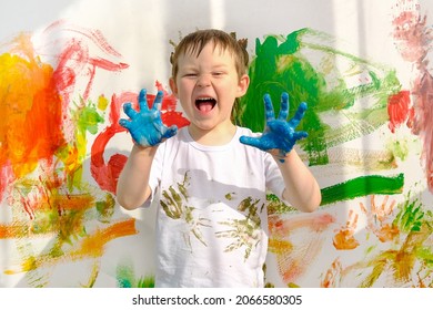 a boy in a white T-shirt, stained with paint, and dirty blue hands, stands against the background of a multi-colored wall. The child painted with his hands. A funny, lovable boy, looks into the frame.