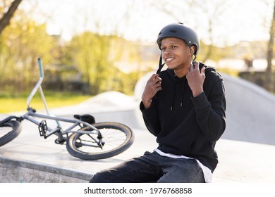 A Boy Who Has Fallen Off Bike, A Bmx, And Is Sitting On A Concrete Ramp, Gets Ready To Ride Back On The Track With Friends, Puts Helmet On His Head And Fastens It Under Neck