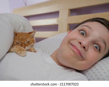 A Boy With Wide Open Eyes Lies In Bed, Smiling. The Boy And The Cat Lie In Bed. Emotions Of Happiness And Joy. Live Style