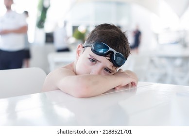 A Boy Without A T-shirt Is Sitting In A Cafe On Vacation And Waiting For A Waiter, Sad. Swimming Goggles On His Head