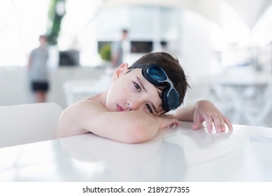 A Boy Without A T-shirt Is Sitting In A Cafe On Vacation And Waiting For A Waiter, Sad. Swimming Goggles On His Head