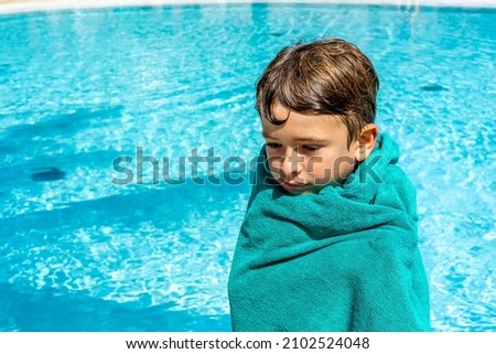 Boy wrapped in towel feeling cold after swimming standing next to pool. High quality photo
