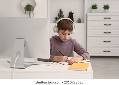 Boy writing in notepad while using computer and headphones at desk in room. Home workplace - Shutterstock ID 2310728585