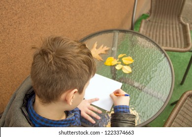 Boy is writing something. Boy is doing homework outdoors. Boy is drawing on paper or writing a letter