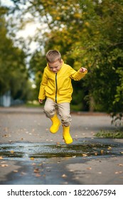 A boy in yellow boots and a yellow jacket is jumping in a puddle.
