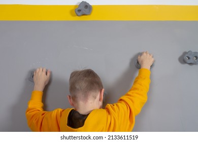 A Boy In A Yellow Hoodie Climbs A Gray Wall On Rock Climbing Stones, A Child Learns Rock Climbing At Home.