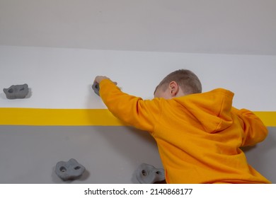 A Boy In A Yellow Hoodie Climbs A Gray Wall On Rock Climbing Stones, A Child Learns Rock Climbing At Home.