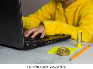 Boy in yellow pajamas sits at the computer, earning on Bitcoin mining. Near the computer is hourglass, pencil and gold bitcoin coin.The child learns to make money. - Powered by Shutterstock