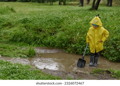 A boy in a yellow raincoat and rubber boots is standing in a stream. In the hands of a shovel. - Powered by Shutterstock