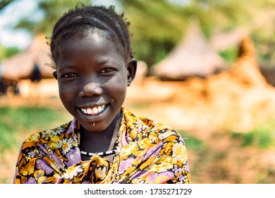BOYA TRIBE, SOUTH SUDAN - MARCH 10, 2020: Girl in traditional colorful outfit and accessory smiling at camera against blurred settlement in South Sudan in Africa