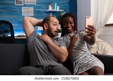 Boyfriend Being Goofy While Joyful Girlfriend Taking Selfie Using Smartphone. Cheerful African American Wife And Caucasian Husband Taking Pictures Together With Phone While Sitting On Sofa In Living