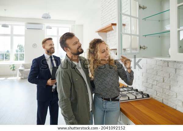 Boyfriend and girlfriend buying house. First time\
buyers or future tenants meeting real estate agent and looking\
around new home. Husband and wife looking at good quality modern\
wooden kitchen\
cabinet