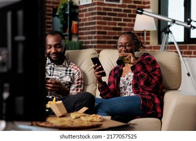 Boyfriend and girlfriend using smartphones and eating takeaway food while they watch movie on television. Takeout pizza, noodles, burger and snacks with bottles of beer watching tv film. - Powered by Shutterstock