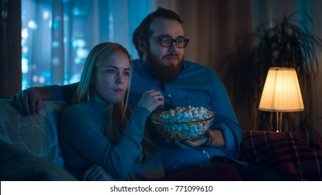 Boyfriend and Girlfriend Watching TV. They Sit on a Sofa in Their Cozy Living Room and Eat Popcorn. It's Evening.