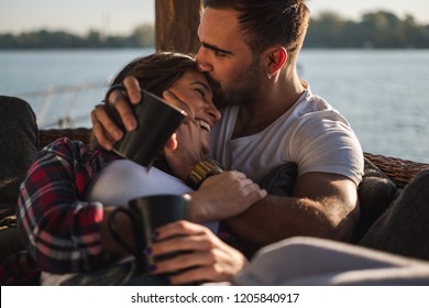 Boyfriend kissing his smiling girlfriend in forehead while she is lying covered with blanket  by the river. Beautiful autumn day