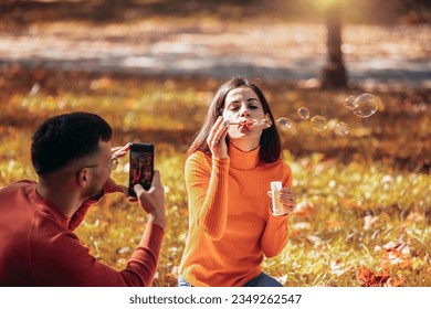 Boyfriend taking a picture of his girlfriend while she blows bubbles. Sunny autumn day in the colorful park - Shutterstock ID 2349262547