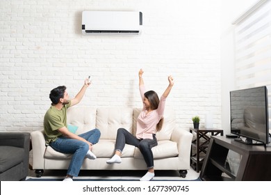 Boyfriend turning on air conditioner while cheerful girlfriend sitting with arms raised on sofa at home - Shutterstock ID 1677390745