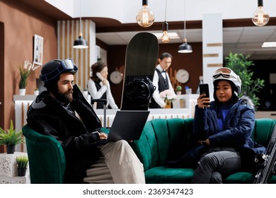 Boyfriend using laptop and girlfriend with mobile phone enjoying digital communication. Wintersports enthusiasts in hotel lobby for ski and snowboarding fun waiting with their smart devices. - Powered by Shutterstock