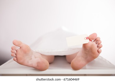 Boys Body Or Cadaver Lying On A Mortuary Slab Covered With A White Sheet With A Blank Tag Tied To His Toe Awaiting Identification, View From The Bare Feet