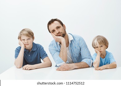 Boys feeling bored and upset. Portrait of tired funny european family of sons and dad sitting at table, leaning heads on palms and staring indifferent at camera, being careless while mom scolding them