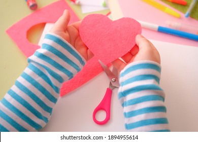 Сhild's Boy's Girl's Hand Holding Heart Cut Out Of Felt. Children's Creativity - Drawings And Crafts  For Valentines Day Or Mother's Day Or Father's Day. Top View.Colored Paper, Pens, Scissors Table