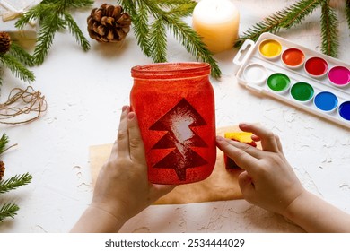 the boy's hands make a candlestick from a glass jar painted with red paint with a silhouette of a Christmas tree, step by step instructions, step 6 - Powered by Shutterstock