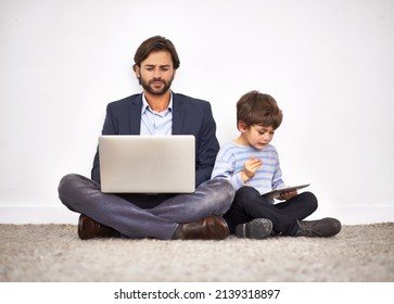 Boys love their digital toys. A father and son sitting on the floor against a wall with a laptop and digital tablet.