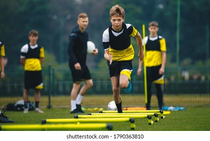 Boys On Football Training Session With Two Young Coaches. Junior Level Soccer Player Running And Doing Skipping Exercise With Hurdles During Practice Session. Soccer Drills With Hurdles Agility