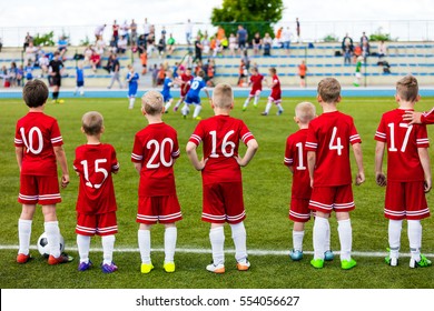Boys Play Soccer Match. Children Sport Team. Youth Sports Team Together. Football Soccer Game For Children. Kids Soccer Players On Bench Watching Tournament Game With Coach. Stadium In The Background