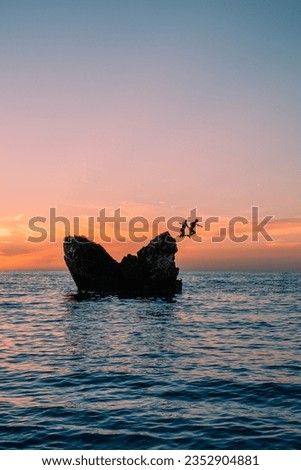 Brace couple jumps off a rock into the ocean during cloudy sunset on the island of Lefkada in summer Greece Europe on their vacation, having fun upright format