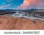 Braided rivers flow through a reddish brown barren landscape in Iceland, with rolling hills and mountains under a vibrant pink and blue sky.