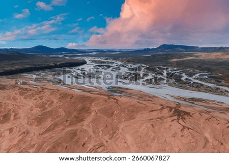 Braided rivers flow through a reddish brown barren landscape in Iceland, with rolling hills and mountains under a vibrant pink and blue sky.