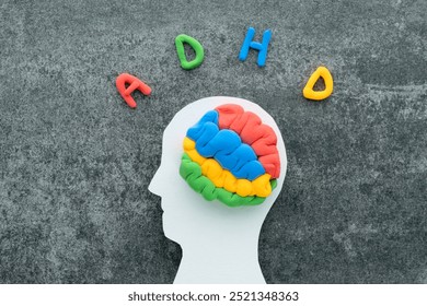 A brain model crafted from colorful materials rests on a white head silhouette, with the letters ADHD on a concrete background. ADHD and learning difficulties awareness and support - Powered by Shutterstock