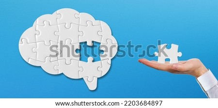 Brain shaped white jigsaw puzzle on blue background. Hand holding a missing piece of the brain puzzle. Mental health and problems with memory. 