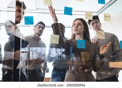 Brainstorm, Presentation And Teamwork Concept. Creative group of diverse professional businesspeople thinking, looking at sticky post-it notes on glass wall discussing new ideas analysing strategy