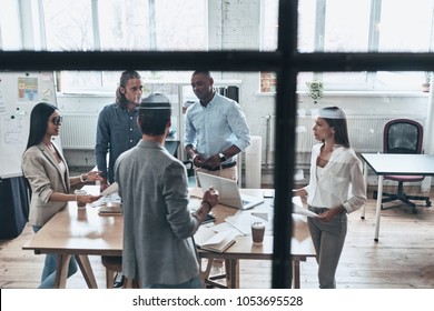 Brainstorming For A New Project. Top View Of Young Modern People In Smart Casual Wear Discussing Business While Standing Behind The Glass Wall In The Board Room