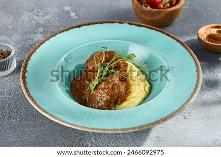 Braised beef cheeks on mashed potatoes in a rustic setting with tomatoes and peppercorn on textured grey background.