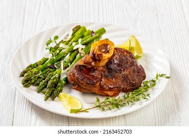 braised osso buco, veal shank steak  with grilled asparagus, lime and swiss chard fresh leaves on white plate on white wooden table