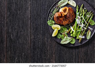 braised osso buco, veal shank steak  with grilled asparagus, lime and swiss chard fresh leaves on black plate on dark wood table, horizontal view from above, flat lay, free space