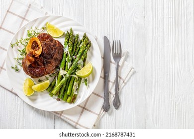 braised osso buco, veal shank steak  with grilled asparagus, lime and swiss chard fresh leaves on white plate on white wood table,  horizontal view from above, flat lay, free space