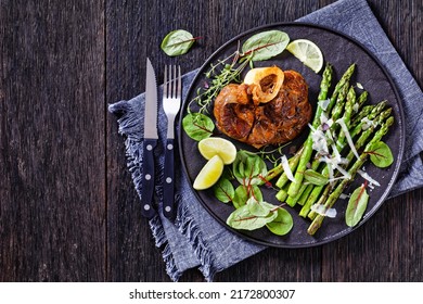 braised osso buco, veal shank steak  with grilled asparagus, lime and swiss chard fresh leaves on black plate on dark wood table, horizontal view from above, flat lay, close-up