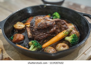 Braised Red Wine Beef Short Ribs Recipe With Vegetables And Bread In Black Pan On Wood Table.