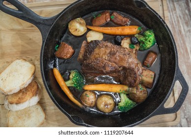 Braised Red Wine Beef Short Ribs Recipe With Vegetables And Bread In Black Pan On Wood Table.