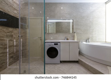 Brand New Bathroom Interior Design. Luxury Modern Bathroom With White Bath Tub, Washing Machine, Walk In Shower With Glass Door.